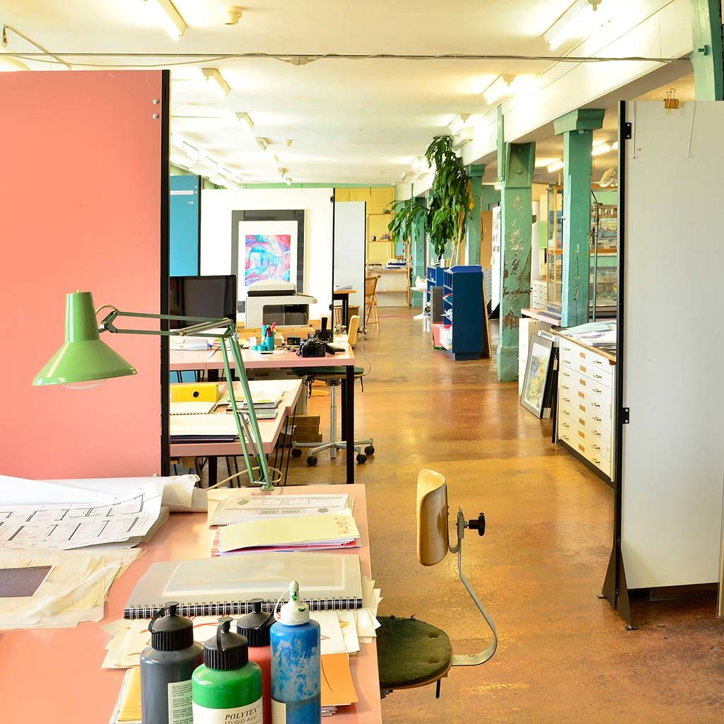 View down the center of an architecture office. A desk with paints and a swing-arm lamp is in the foreground. Other desks are in the background