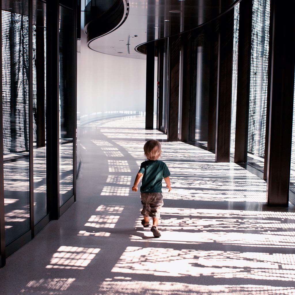 A young boy walks down an interior hallway. The floor is filled with shadows from the building's outdoor patio cover.